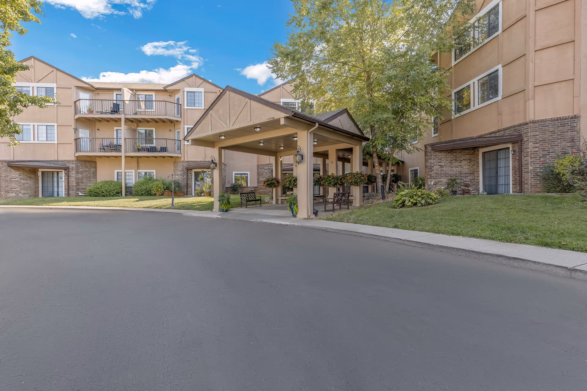 Front entrance of a senior living building with a covered porte-cochère, balconies, benches, and landscaping.