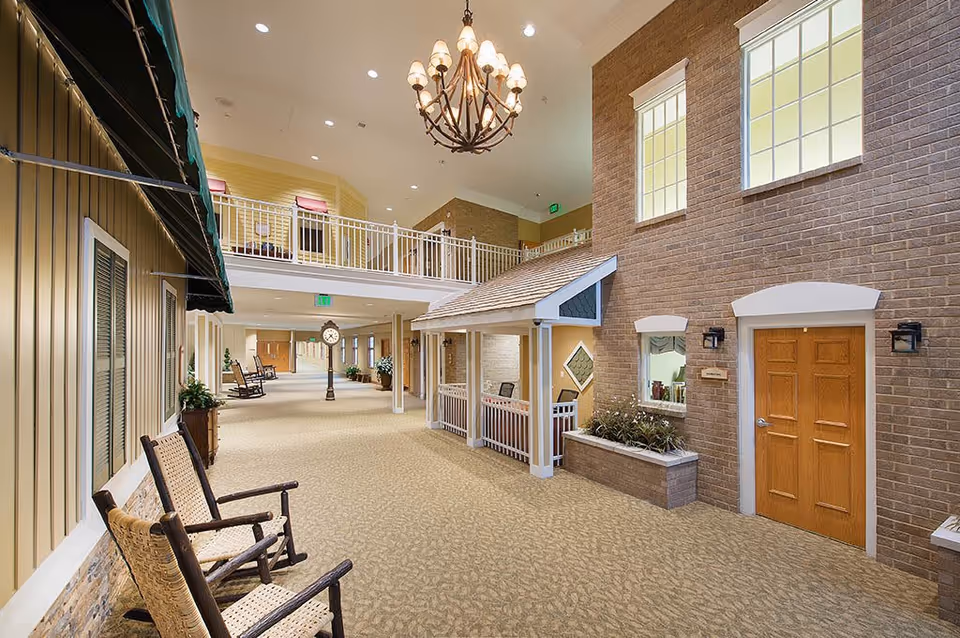 Interior hallway of a senior living facility designed to look like an outdoor street scene with building facades, windows, doors, rocking chairs, plants, and a large chandelier hanging from the ceiling.