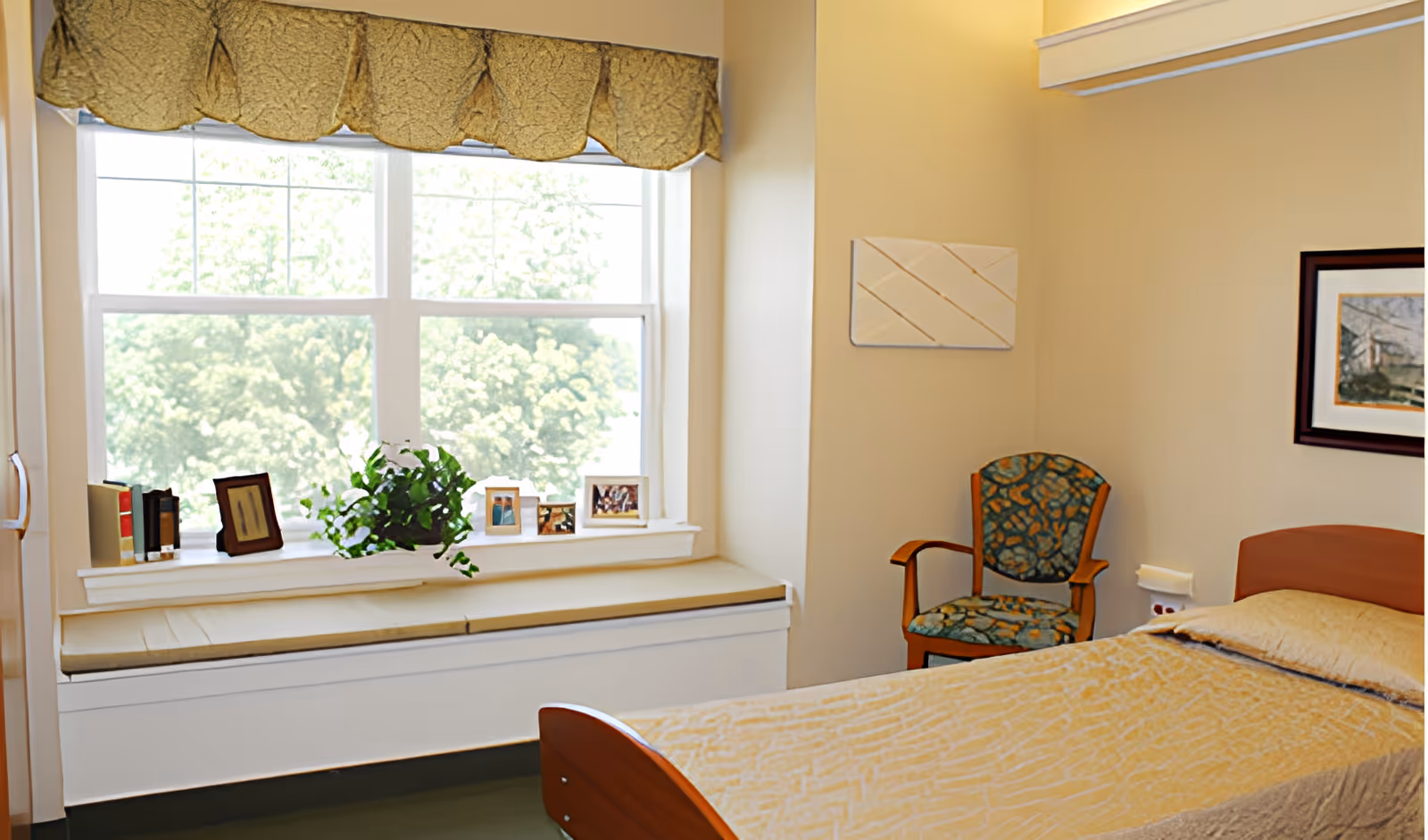 A bright bedroom with a large window featuring a cushioned window seat decorated with framed photos, books, and a potted plant. There is a single bed with a beige bedspread and a wooden headboard, and a patterned armchair next to the bed. The walls are light-colored with minimal artwork.