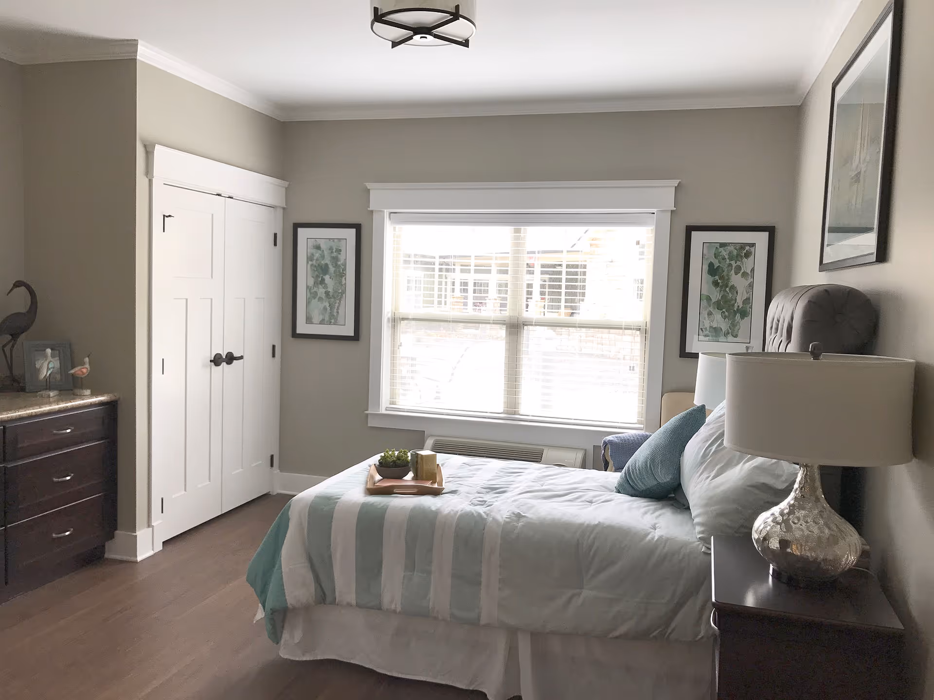 A neatly arranged bedroom with a bed featuring a striped comforter and pillows. There is a nightstand with a decorative lamp beside the bed, a window with blinds letting in natural light, two framed botanical prints on the wall, a dresser with decorative items, and a closed white closet door.