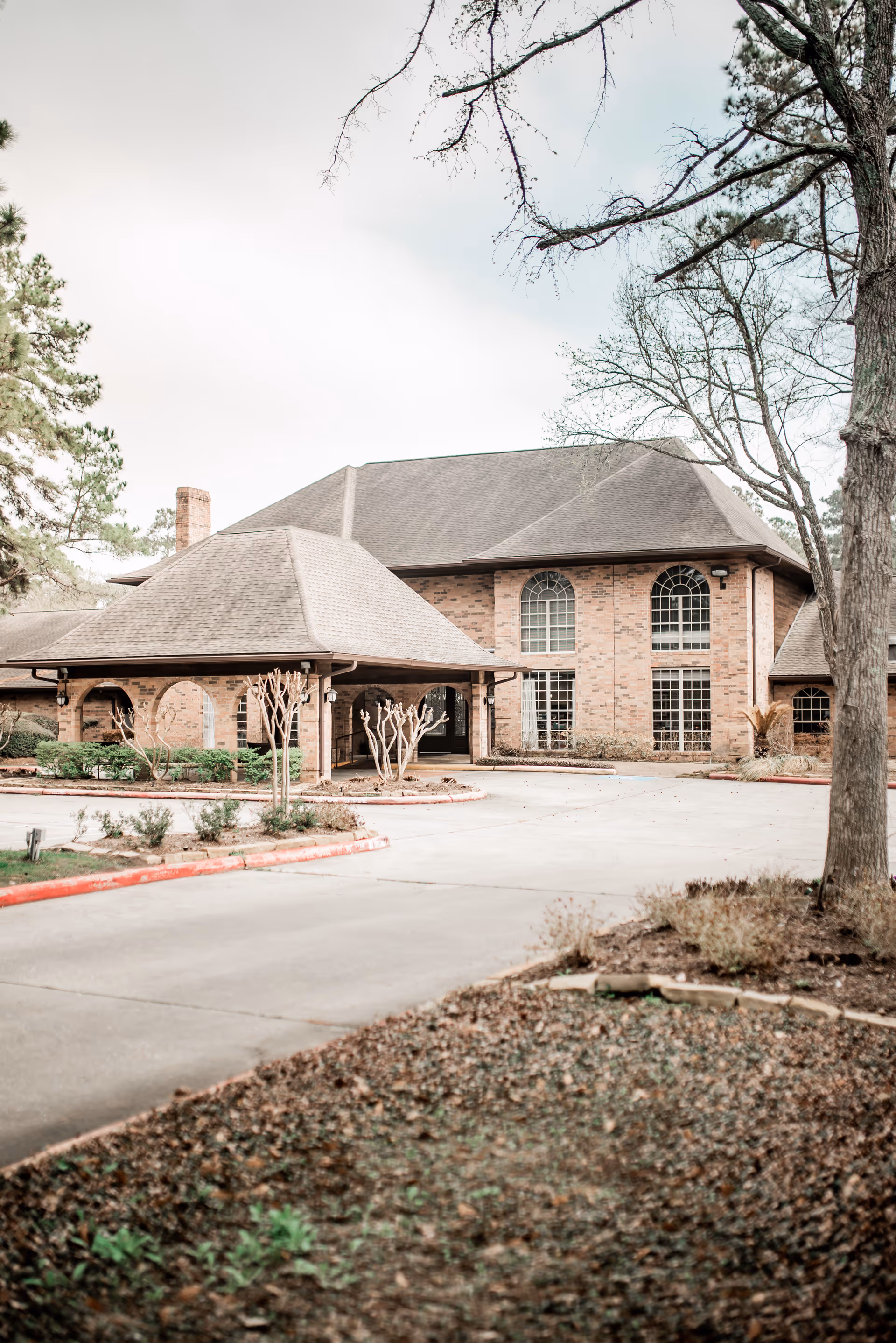 Exterior view of a brick building with large arched windows and a covered entrance driveway, surrounded by trees and landscaping.