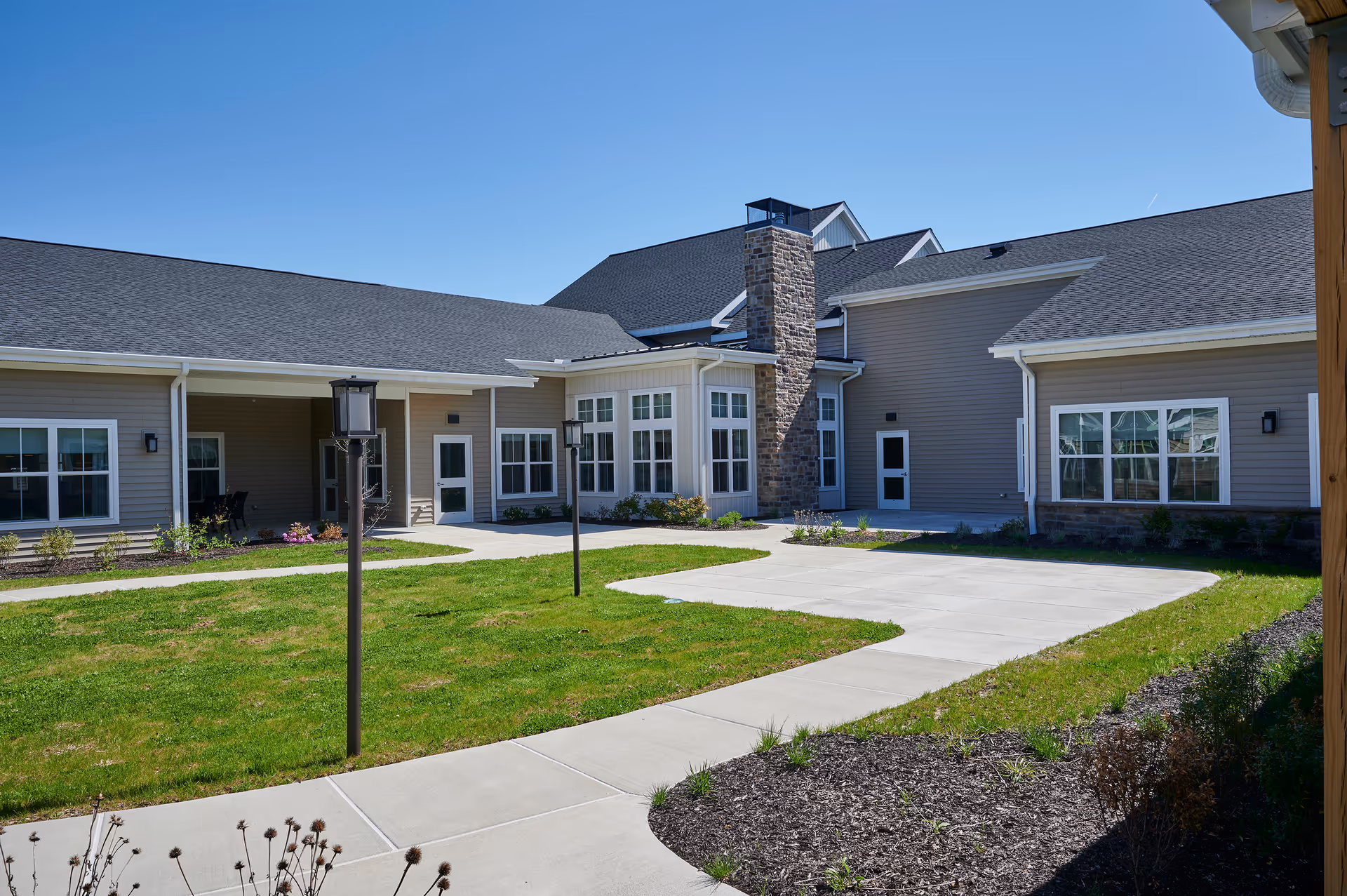 Exterior view of Columbia Cottage of Hanover showing a single-story building with beige siding, multiple windows, a stone chimney, and a paved walkway surrounded by green grass and landscaped areas under a clear blue sky.
