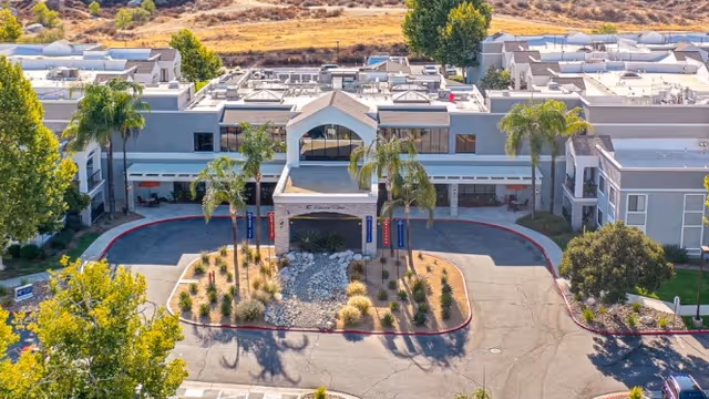Aerial view of the front entrance of Menifee Senior Living showing the porte-cochère, driveway, palm trees, and landscaped roundabout.