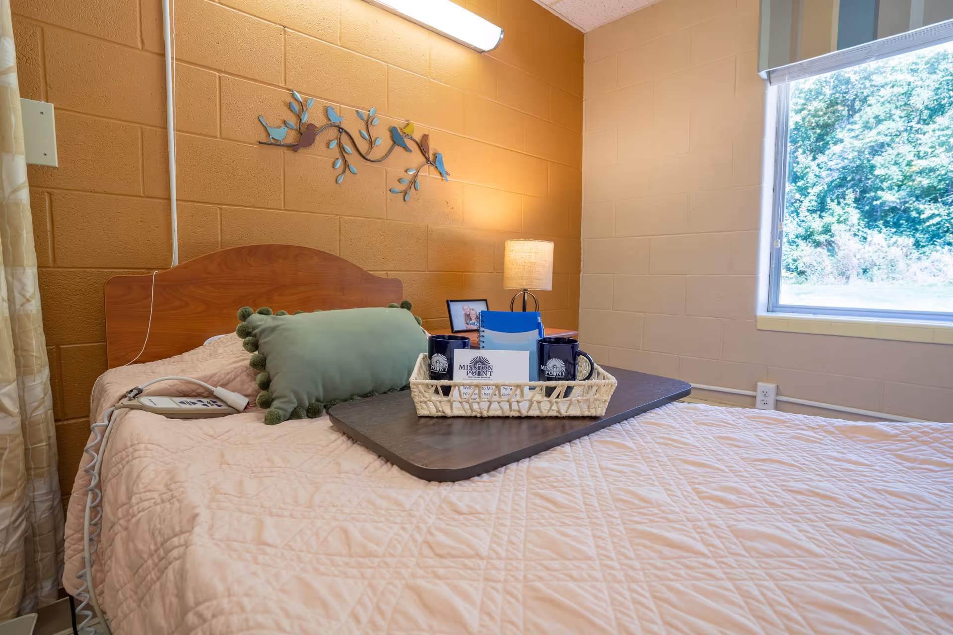 A cozy bedroom with a single bed featuring a light pink quilted bedspread and a green decorative pillow. A wooden headboard is against a mustard yellow brick wall adorned with a metal wall decoration of birds on branches. On the bed is a dark wooden tray holding a wicker basket with two black mugs and some papers. A small lamp and a framed photo sit on a bedside table next to the bed. A window on the right side shows greenery outside.