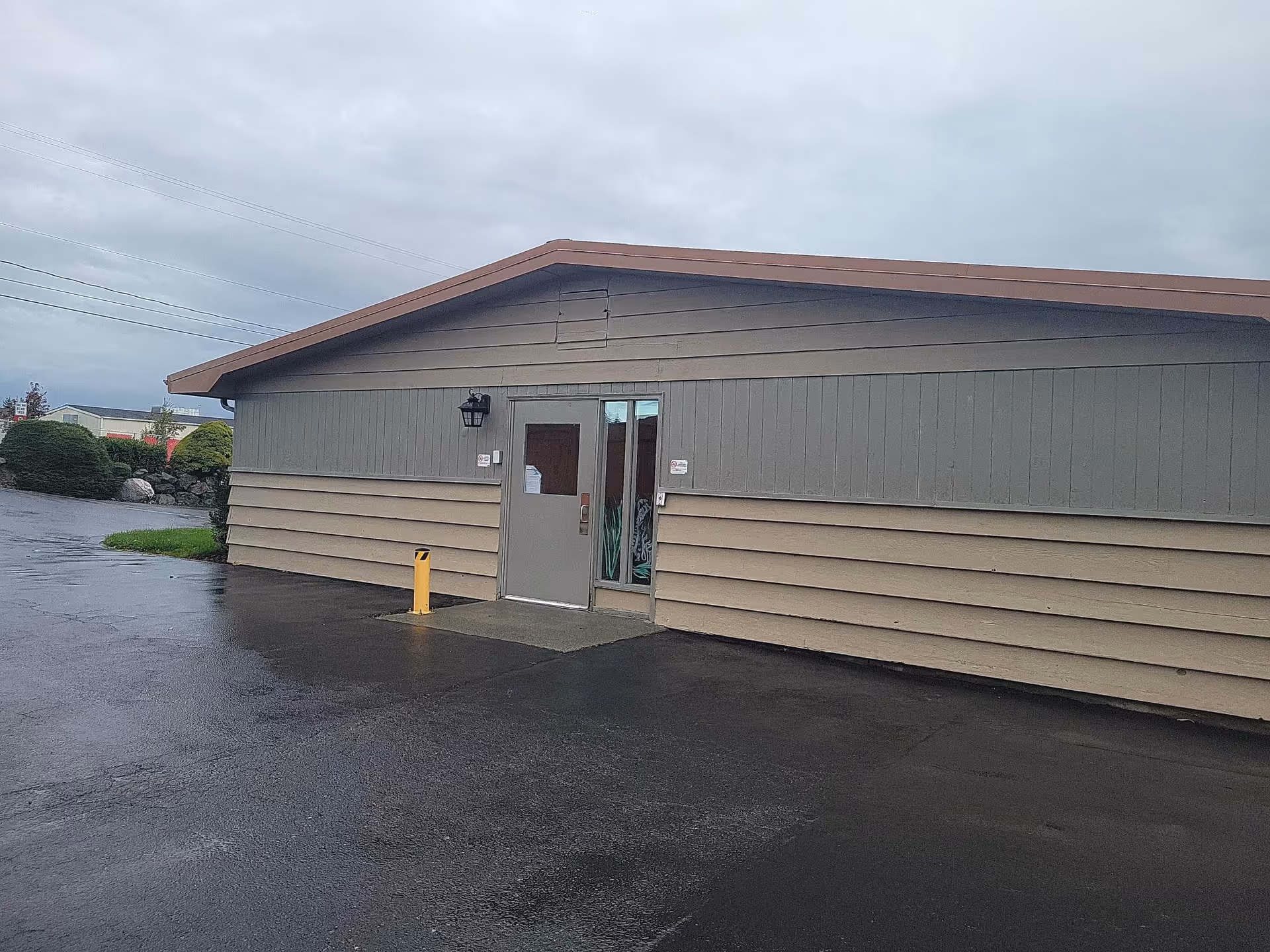 Exterior view of a single-story building with beige and gray siding, a brown roof, and a gray door with glass panels. The ground is wet, indicating recent rain, and there are bushes and other buildings visible in the background under a cloudy sky.