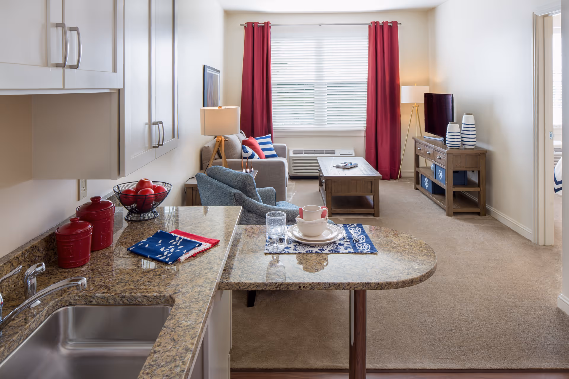 Open-plan living area with a kitchen counter in the foreground, a blue armchair, coffee table and TV stand, and red curtains over a window.