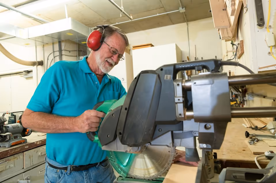 An older man wearing a turquoise polo shirt and red ear protection is operating a large power saw in a workshop. He is smiling and appears focused on cutting a piece of wood. The workshop has various tools and equipment visible in the background.