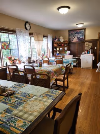 A cozy dining room with wooden floors and multiple tables covered with colorful tablecloths. Several wooden chairs are arranged around the tables. Large windows with sheer curtains allow natural light to fill the room. Shelves with decorative items and a painting are visible on the far wall.
