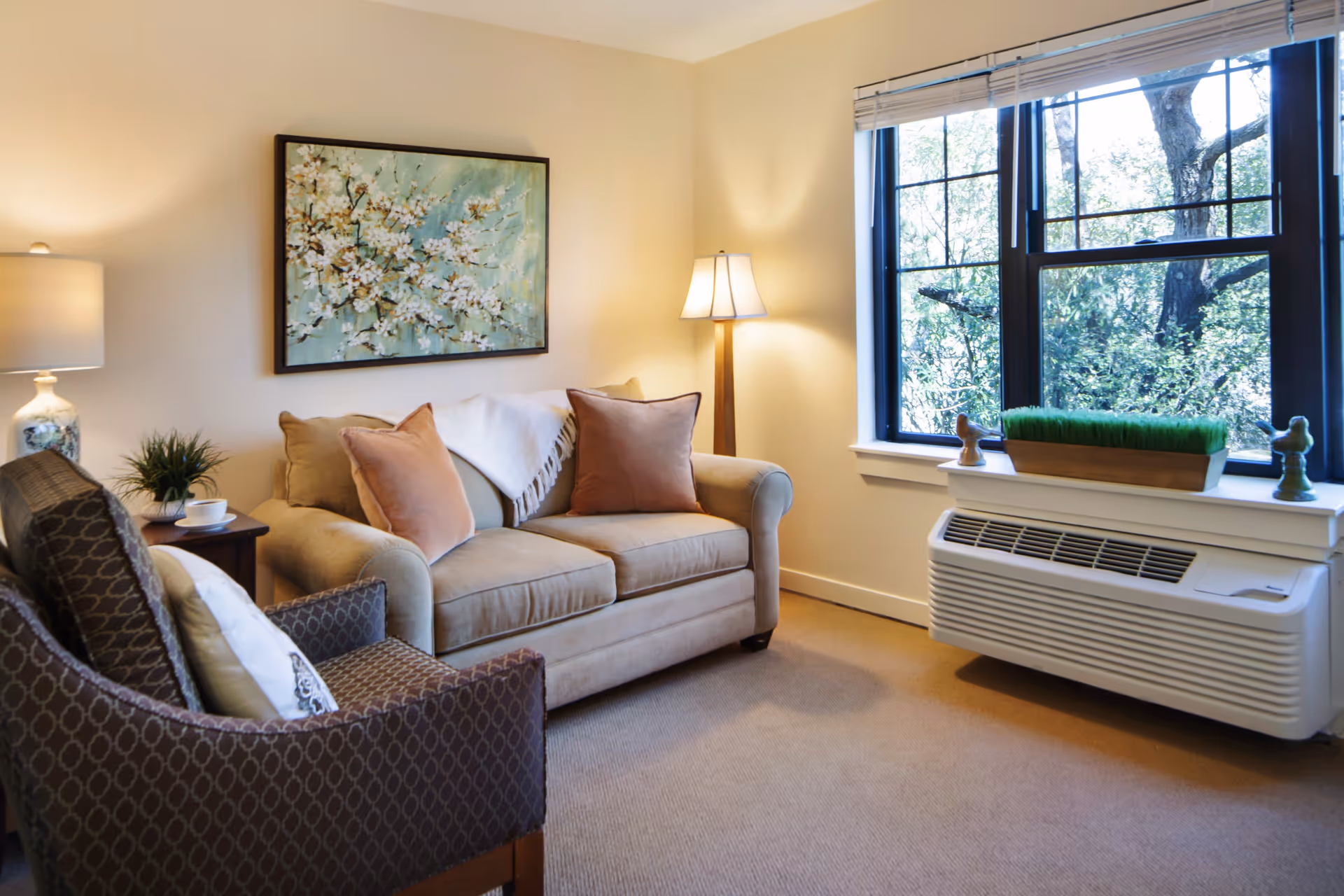 A cozy living room with a beige loveseat adorned with two pinkish-brown pillows and a white throw blanket. Next to it is a patterned armchair with a white pillow. A small wooden side table holds a lamp, a small plant, and a cup. On the wall above the loveseat is a framed painting of white blossoms on a green background. A large window with white blinds lets in natural light, and below it is a white air conditioning unit with decorative items on the windowsill. The room has beige walls and carpeted flooring.