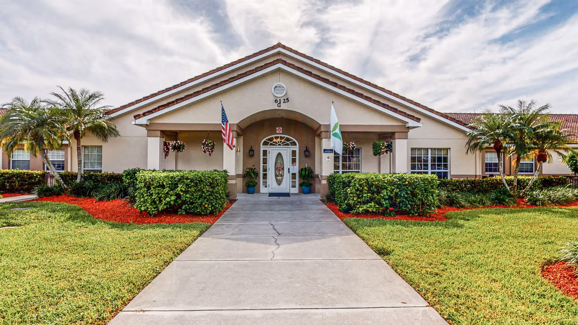 Front exterior view of a single-story senior living community building with a tiled roof, an entrance walkway, two flags on either side of the entrance, hanging flower baskets, palm trees, and well-maintained landscaping with green grass and red mulch.