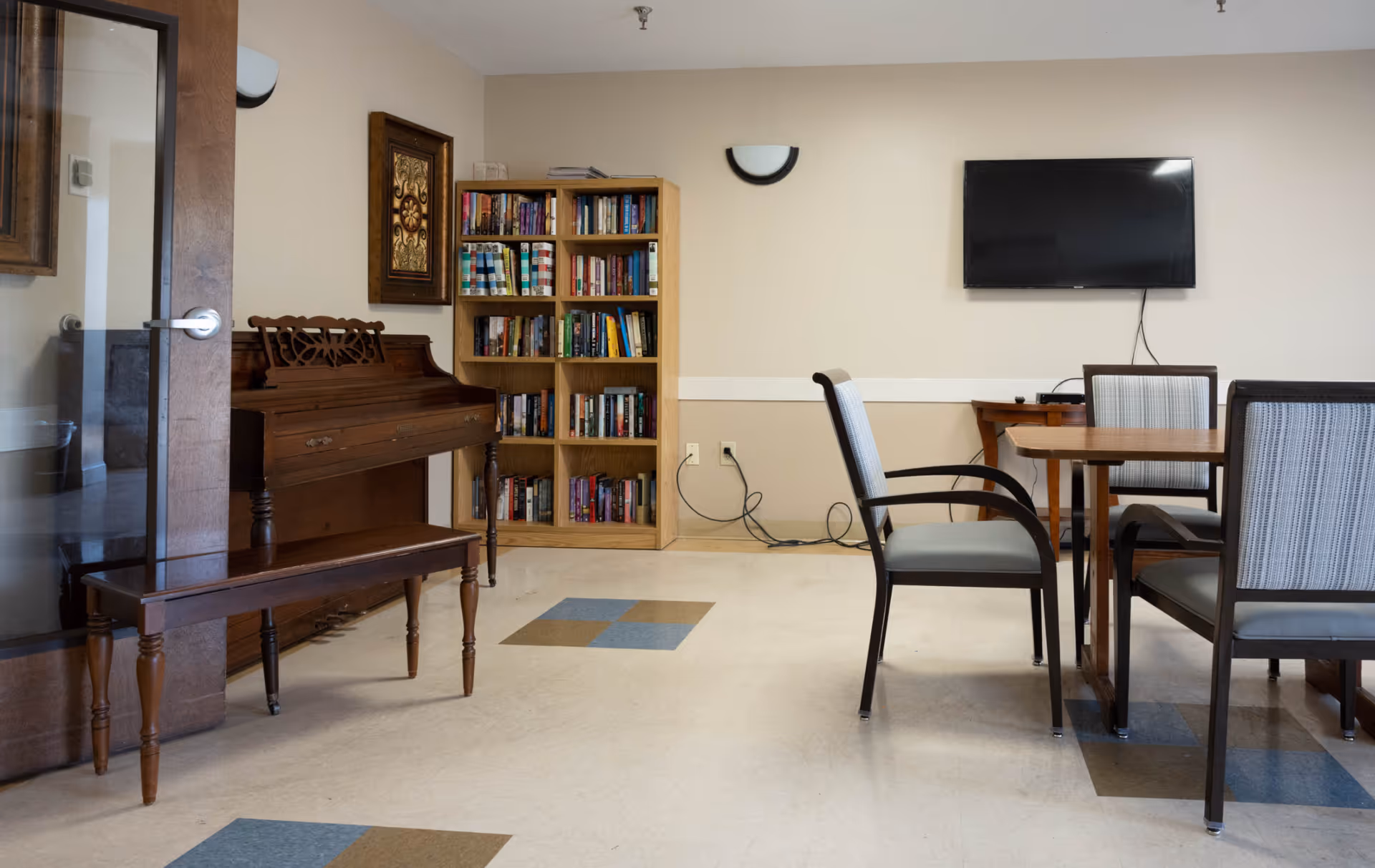 A cozy room in Four Oaks Senior Living featuring a wooden piano with a matching bench, a bookshelf filled with books, a wall-mounted flat screen TV, and a wooden table surrounded by four cushioned chairs. The room has beige walls and a tiled floor with blue and brown square patterns.