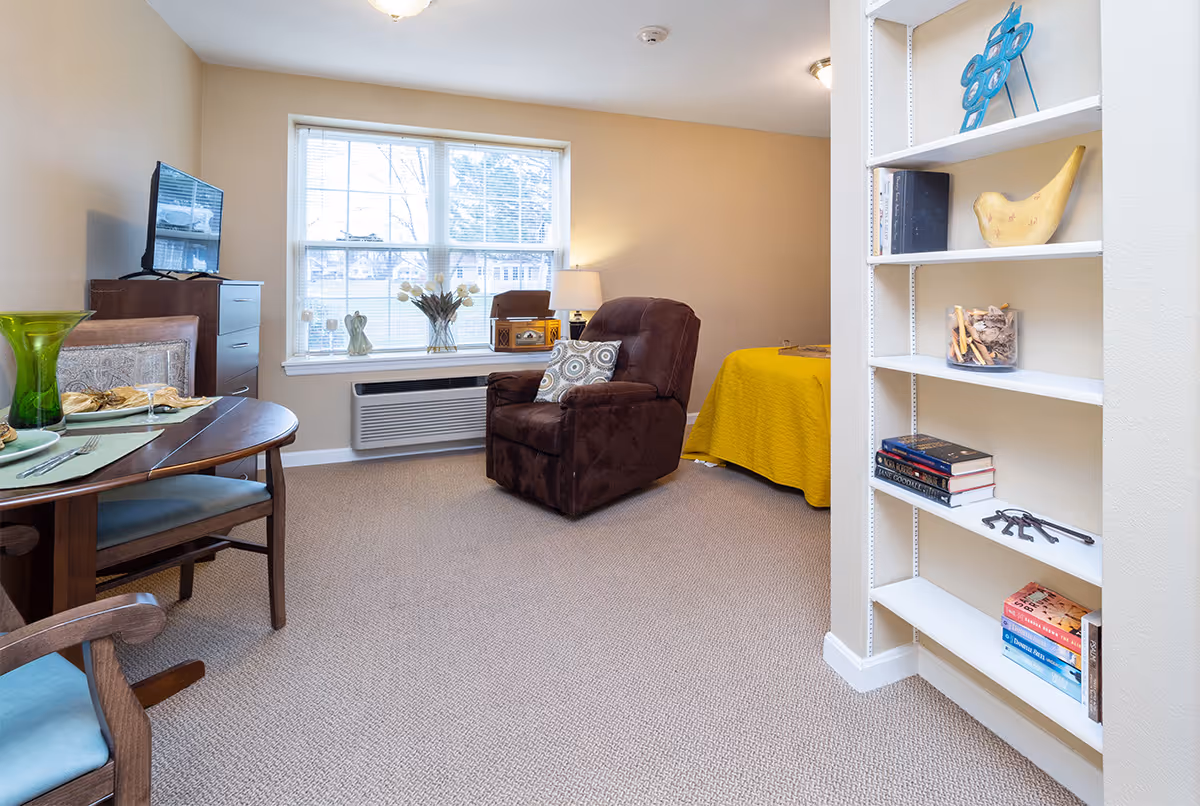 A cozy living space with a brown recliner chair, a yellow bedspread on a bed, a wooden dining table with two chairs, a TV on a dresser, and a white shelving unit with books and decorative items. A large window lets in natural light.