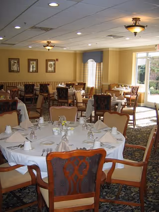 A dining room with round tables covered in white tablecloths, each set with folded napkins, cups, glasses, and silverware. The room has upholstered wooden chairs, framed artwork on the walls, and large windows letting in natural light.
