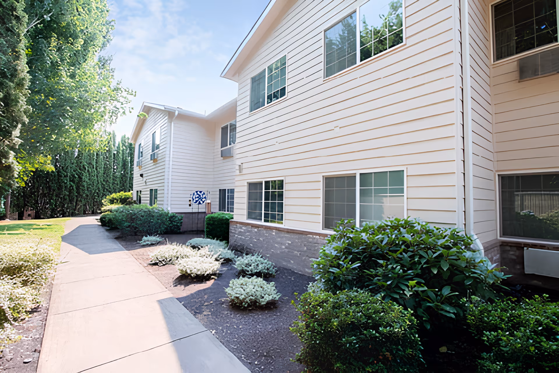 Exterior view of a two-story senior living facility building with white siding and multiple windows. A paved walkway runs alongside the building, bordered by neatly trimmed bushes and small plants. Tall trees and greenery are visible in the background under a clear sky.