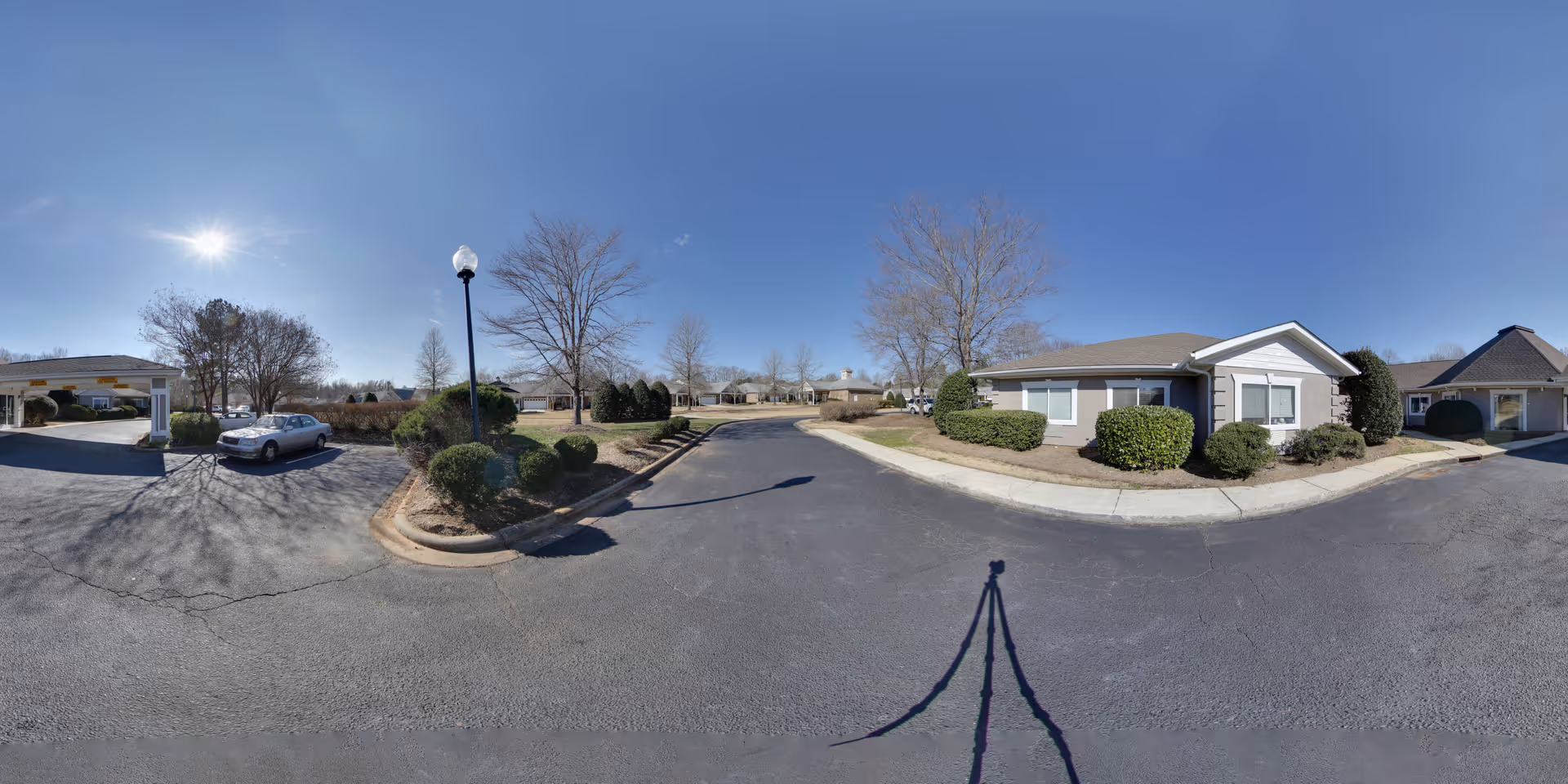Wide outdoor view of a senior living facility parking area with a few single-story buildings, leafless trees, bushes, a streetlamp, and a parked car under a clear blue sky.