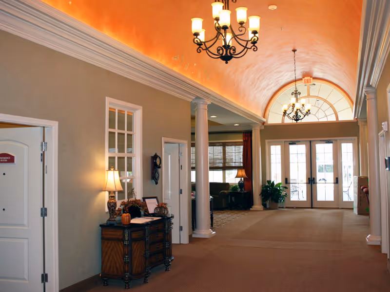 Interior hallway of a senior living facility with beige walls, a carpeted floor, decorative columns, and two chandeliers hanging from a vaulted ceiling with warm lighting. There is a wooden console table with a lamp and decorative items on the left side, and double glass doors at the far end leading outside. A seating area with chairs and a lamp is visible through an open doorway.