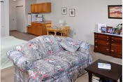 Cozy studio living area showing a floral loveseat, coffee table, dresser, small kitchenette and dining table with a bed partially visible in the background.