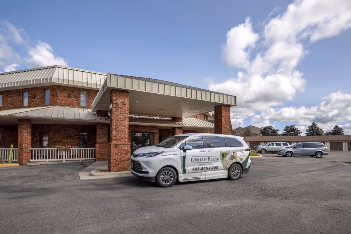 Front entrance of a brick senior living building with a covered porte-cochere and a branded Derian Place van parked in front.