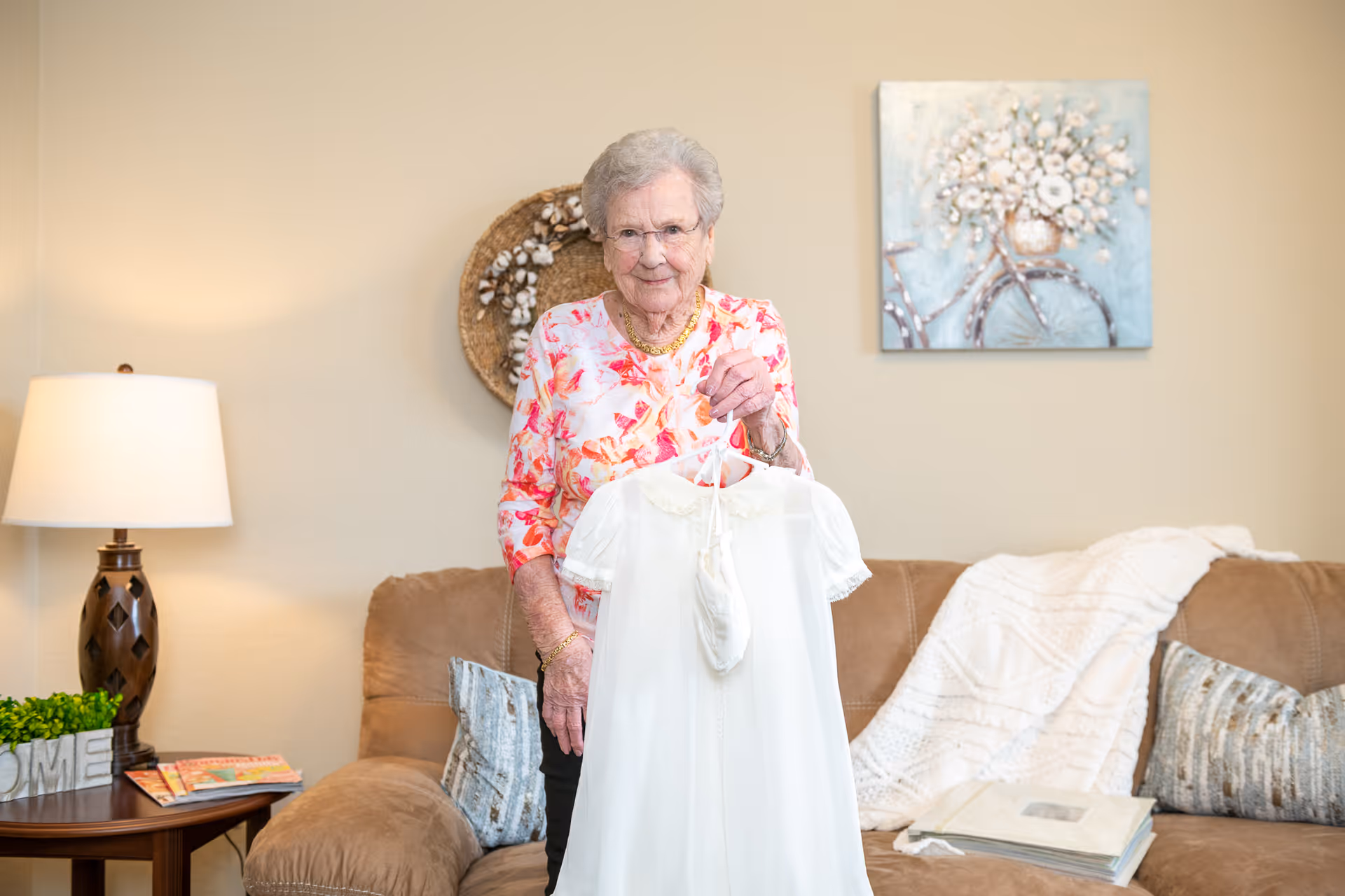 An elderly woman in a floral blouse stands in a living room holding a white dress on a hanger.
