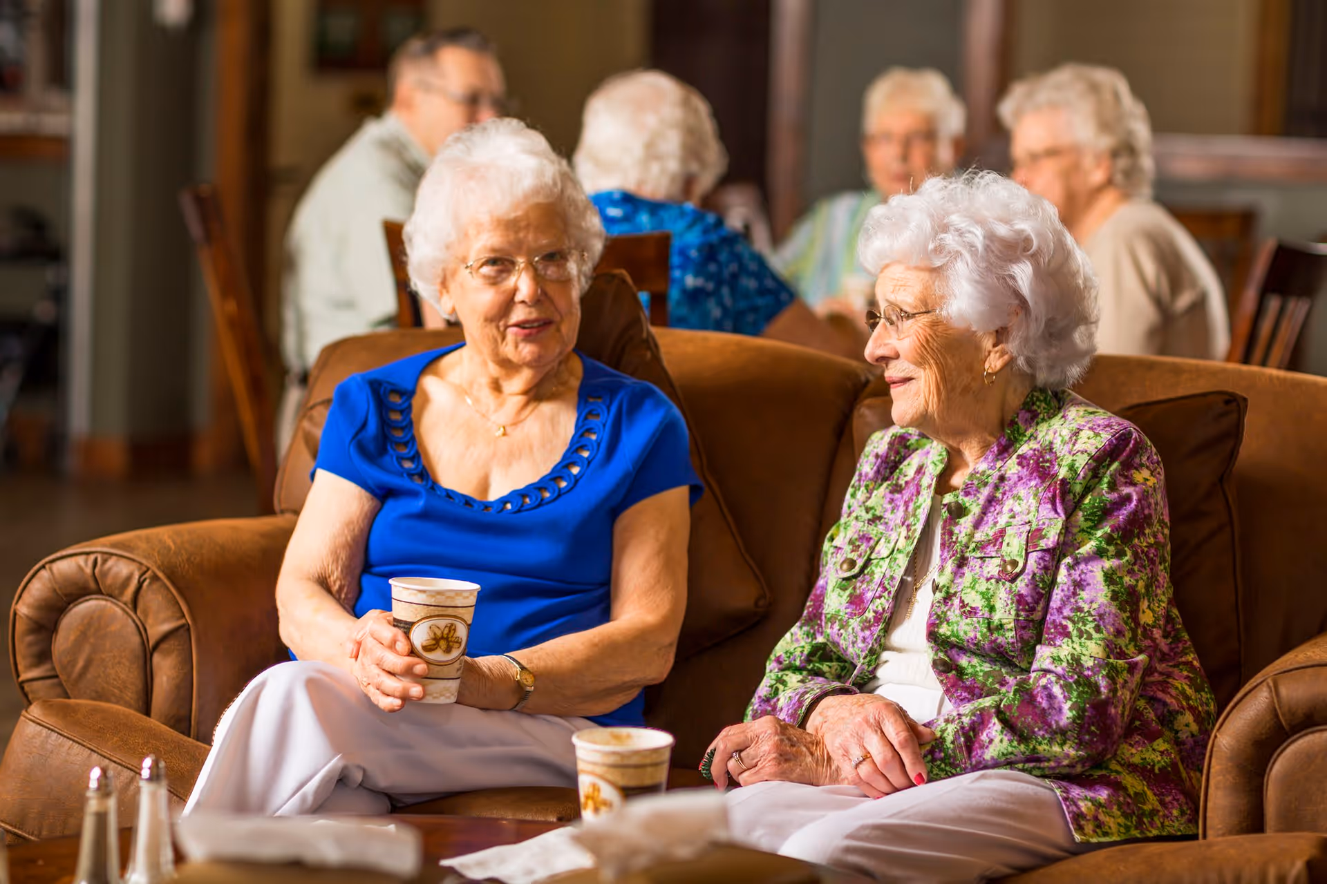 Two elderly women sitting on a brown leather couch in a communal living area, engaged in conversation while holding disposable coffee cups. In the background, other elderly individuals are seated and interacting in a warmly lit room.