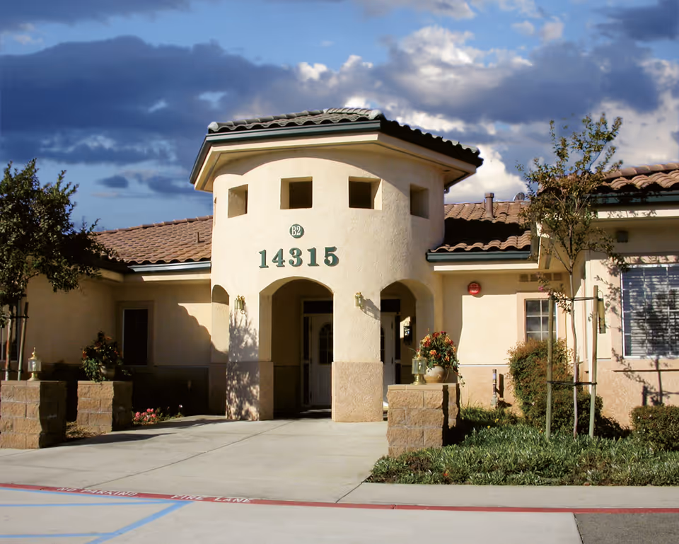 A tan stucco senior living facility entrance with arched doorways and landscaping.