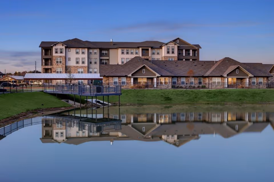 Front exterior of a multi-story senior living building reflected in a calm pond at dusk.