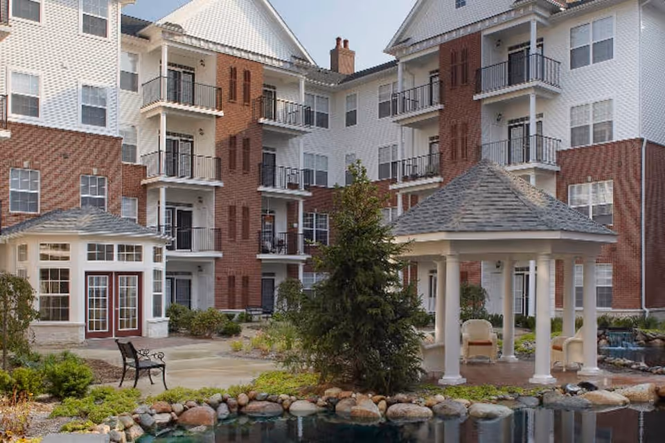 Outdoor courtyard area of a senior living facility with a pond surrounded by rocks, a small gazebo with white columns and a gray roof, benches, and a multi-story building with balconies and large windows in the background.