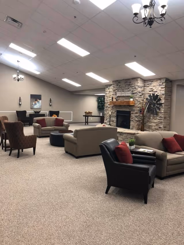 A spacious living room area in an assisted living facility featuring multiple sofas and armchairs with red cushions arranged around a stone fireplace. The room has beige carpeting, neutral-colored walls, ceiling lights, and decorative elements including a wall-mounted windmill and framed artwork.