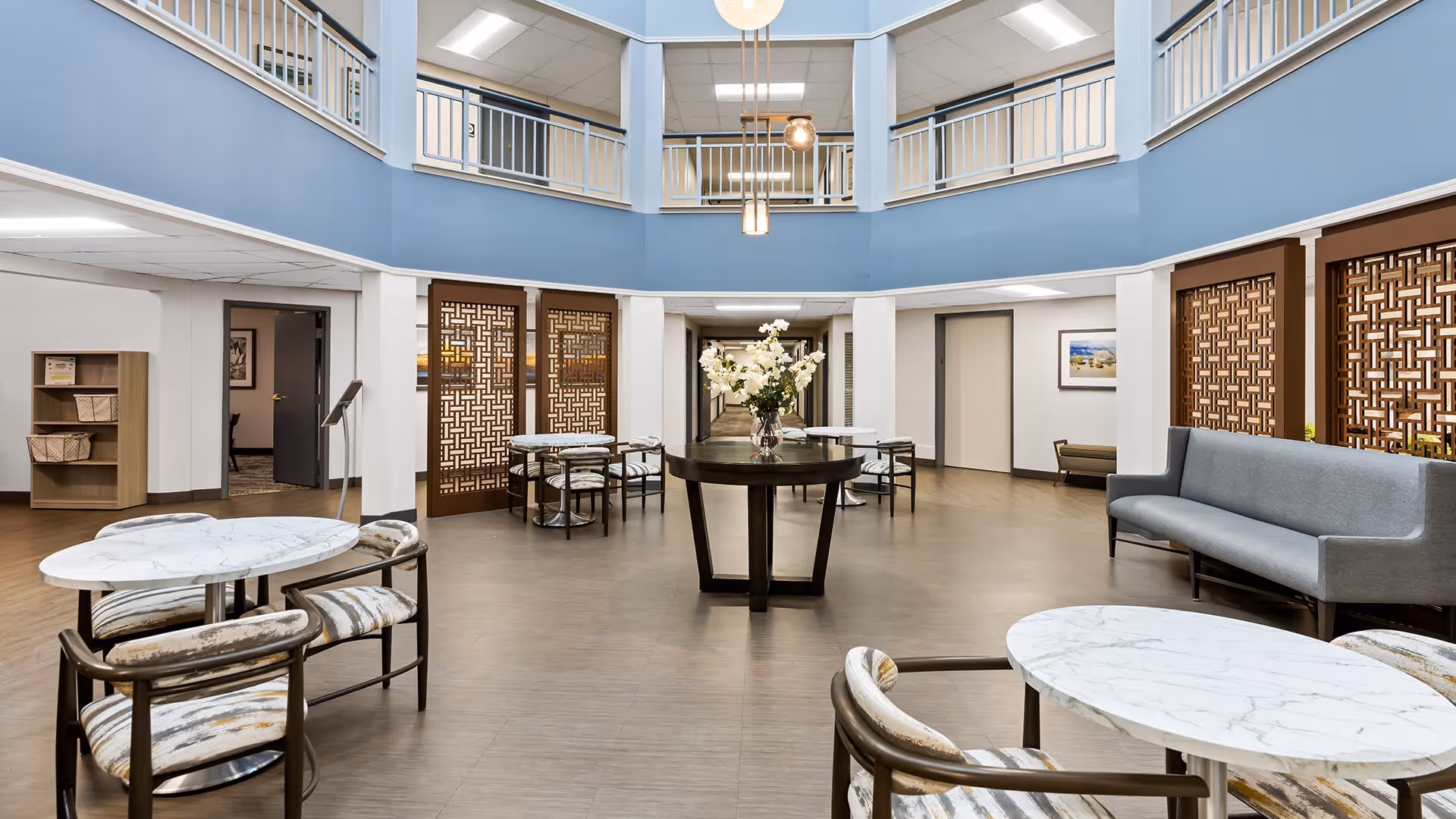 A spacious, modern common area with a high ceiling and a second-floor balcony railing. The room features several round tables with marble tops and cushioned chairs, a central dark wood table with a vase of white flowers, a gray upholstered bench, decorative wooden panels, and soft lighting fixtures hanging from the ceiling.