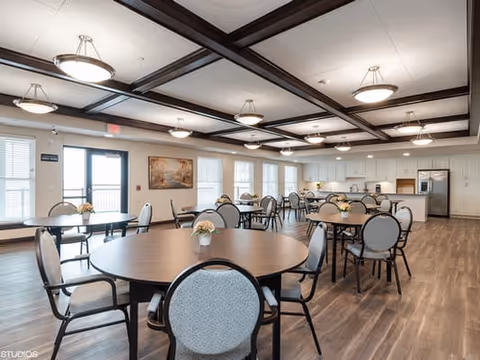 A spacious dining room with multiple round tables and cushioned chairs arranged neatly. The room features wood flooring, a coffered ceiling with dark wooden beams, and modern hanging light fixtures. Large windows along one wall allow natural light to fill the space. In the background, there is a kitchen area with stainless steel appliances and white cabinetry. A framed painting is hung on the wall near the windows.