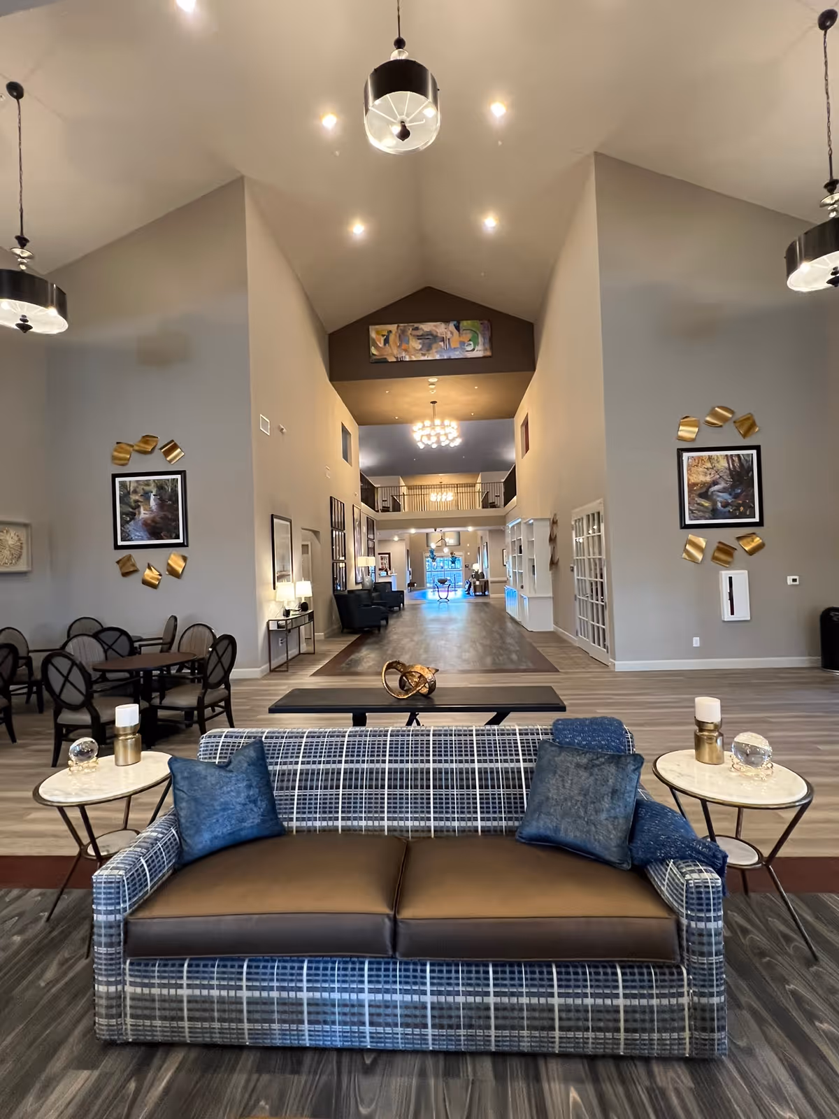 Spacious and well-lit common area in an assisted living facility featuring a plaid sofa with two blue pillows in the foreground, two round side tables with decorative items, multiple round dining tables with chairs on the left, and a long hallway leading to more seating and a balcony area above. The walls are decorated with framed artwork and gold accents, and pendant lights hang from the high ceiling.