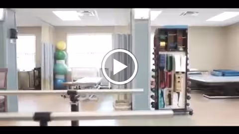 Physical therapy or rehabilitation room with exercise equipment including therapy balls, parallel bars for walking support, and padded treatment tables in a bright, well-lit space with large windows.
