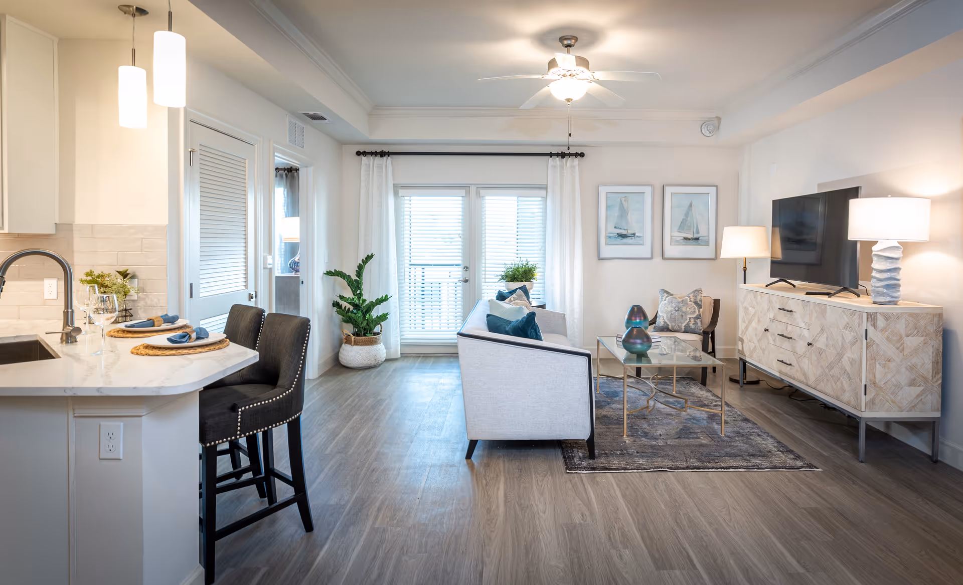 Bright and modern living room with a light gray sofa adorned with blue and patterned pillows, a glass coffee table with decorative items, a wooden TV console with a flat-screen TV, two table lamps, and two framed sailboat paintings on the wall. Adjacent to the living area is a kitchen counter with two black cushioned bar stools, place settings, and hanging pendant lights. The room has wood flooring, white walls, a ceiling fan with light, and a large window with white curtains and blinds.