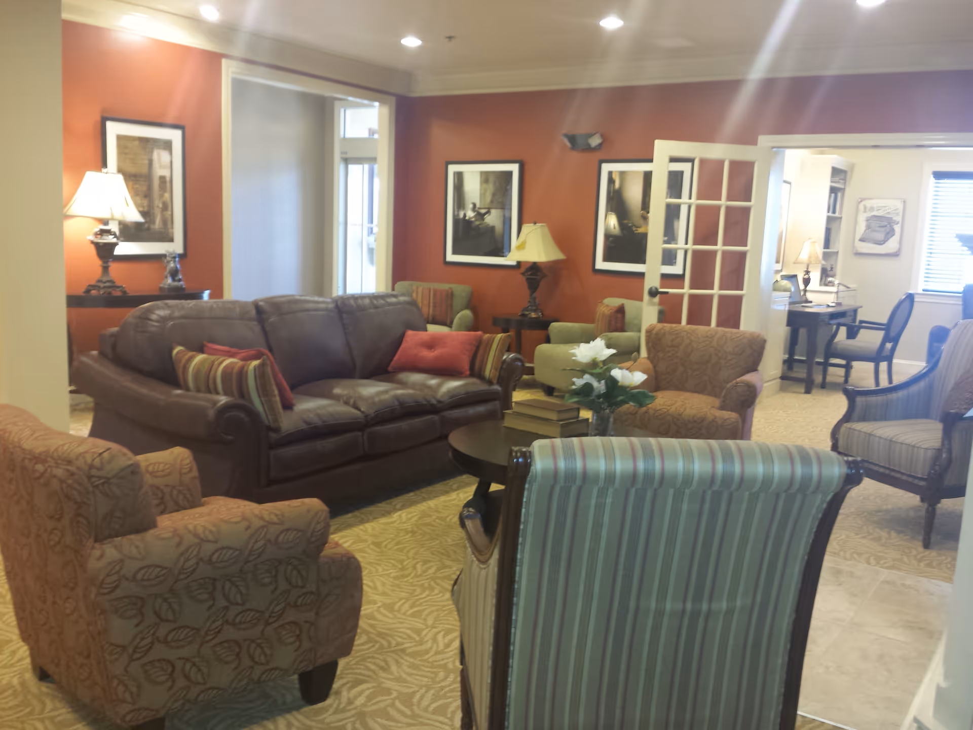 A cozy senior living room with a brown leather sofa, several patterned armchairs, a round coffee table with books and a flower vase, warm orange walls with framed artwork, and lamps providing soft lighting. A doorway with glass-paneled double doors leads to another room with a desk and chair.
