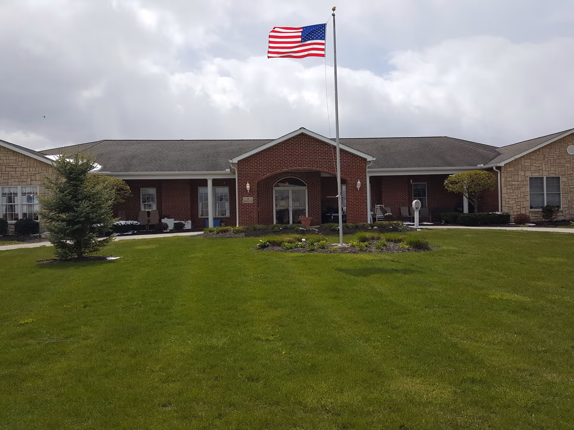 Front exterior view of a single-story senior living facility building with a mix of brick and stone facade, a central entrance with a covered porch, an American flag on a flagpole in the foreground, and a well-maintained green lawn with small trees and shrubs.