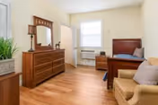 A bright and clean assisted living bedroom featuring a wooden bed with pillows, a matching wooden dresser with a mirror, a nightstand, a beige armchair with cushions, a window with white blinds, and a small air conditioning unit below the window. The room has light-colored walls and wood flooring.