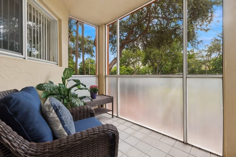A cozy screened-in patio with a wicker chair featuring blue and gray cushions, a small wicker side table with a potted plant, and a view of trees and greenery outside.