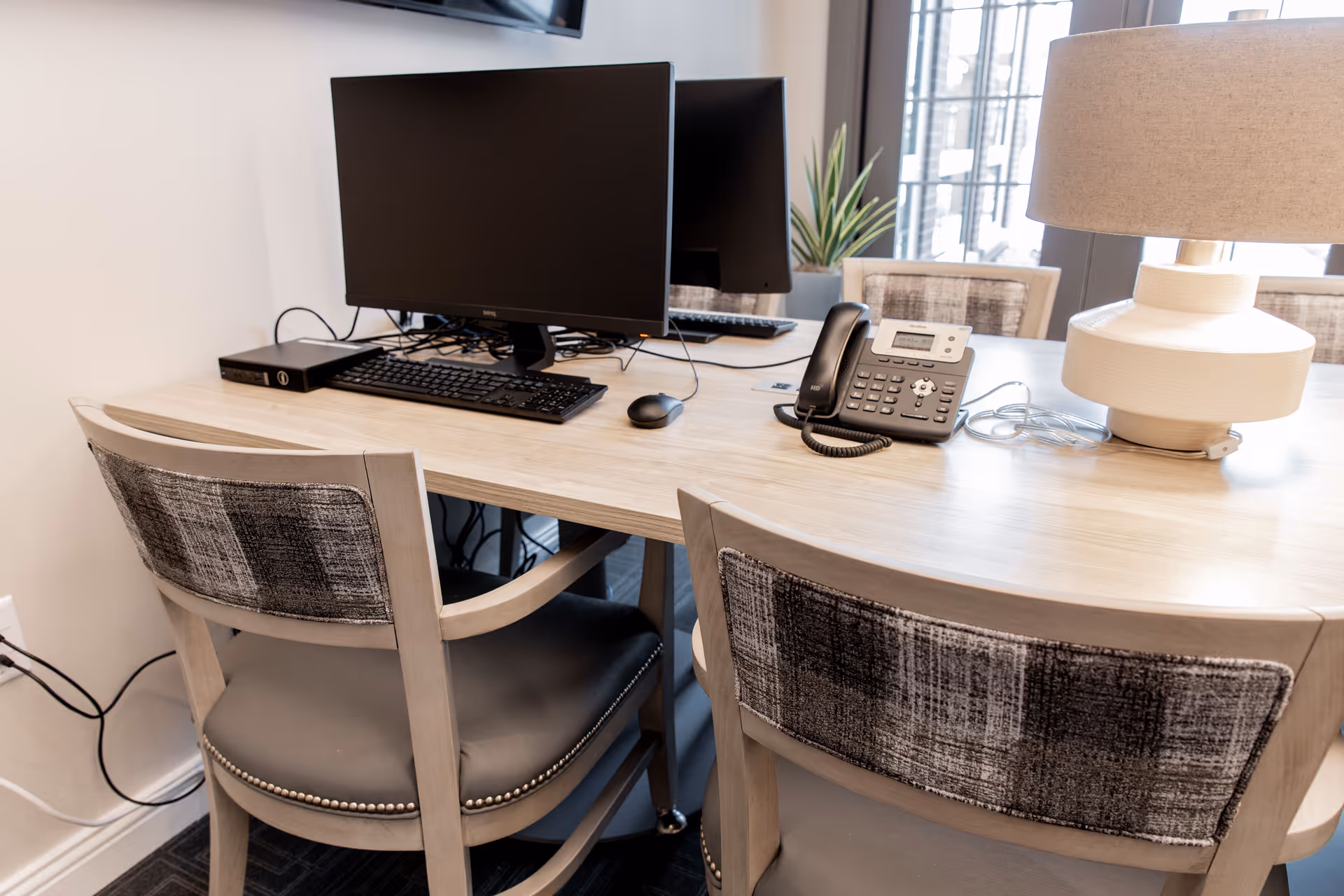 A light wood desk with two chairs featuring gray patterned backs and cushioned seats. On the desk are two computer monitors, a keyboard, a mouse, a telephone, and a beige table lamp. A window with natural light and a small plant are visible in the background.