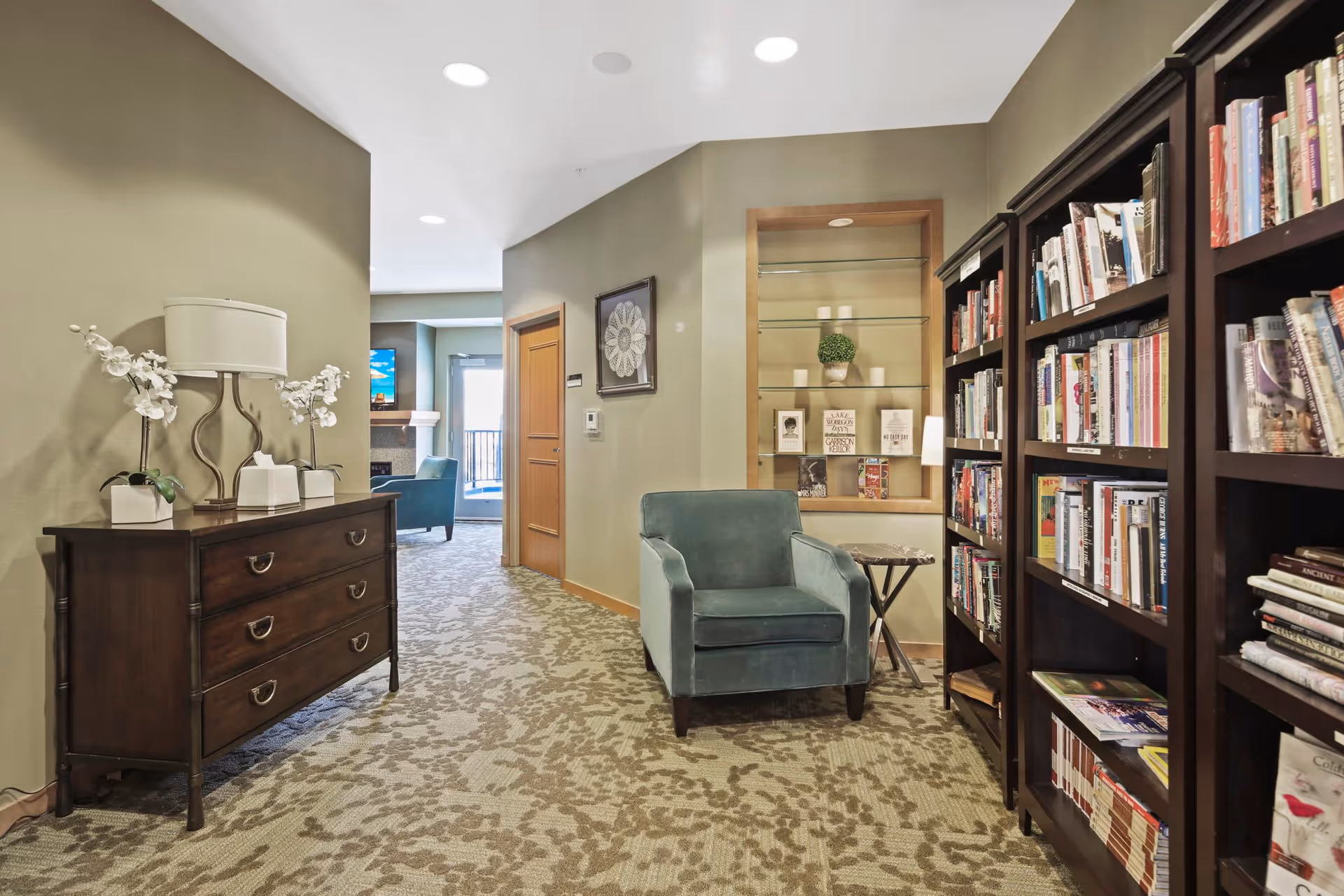 Interior view of a senior living facility hallway with a patterned carpet. On the left, there is a dark wooden chest of drawers with a lamp and white orchid plants on top. On the right, there are tall bookshelves filled with books and a blue armchair next to a small side table. In the background, there is a seating area with a fireplace and a television mounted above it.