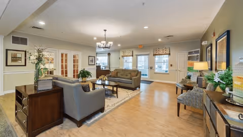 A spacious and well-lit living room area in Sycamore Manor featuring two gray sofas, a wooden coffee table on a patterned rug, decorative plants, framed artwork on the walls, and a chandelier hanging from the ceiling. The room has light-colored walls, wooden flooring, and multiple windows allowing natural light to enter.
