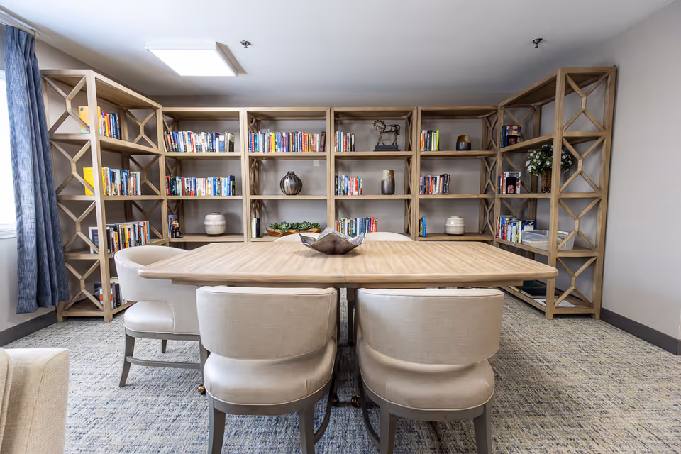 A communal reading room with a wooden table and upholstered chairs facing large wooden bookshelves filled with books and decorative items.
