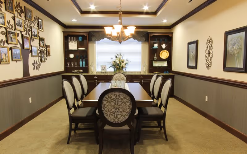 A formal dining room with a rectangular wooden table surrounded by eight upholstered chairs. The room features a chandelier hanging above the table, built-in dark wood shelves and cabinets at the far end, decorative wall art including framed pictures and a metal wall sculpture, and a window with blinds behind a floral centerpiece on the table.