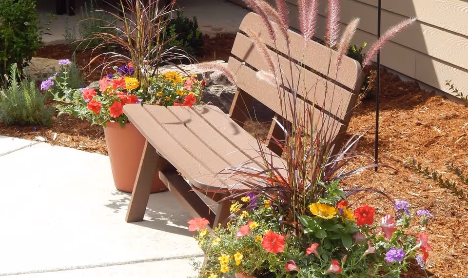 Brown outdoor bench with colorful potted flowers on a sunlit patio beside a building.