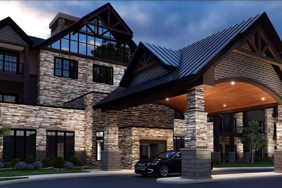Exterior view of a senior living facility named VITALIA Highland Heights during dusk, showcasing a large stone and wood building with a covered entrance where a black car is parked. The building features multiple windows and a peaked roof with architectural details.