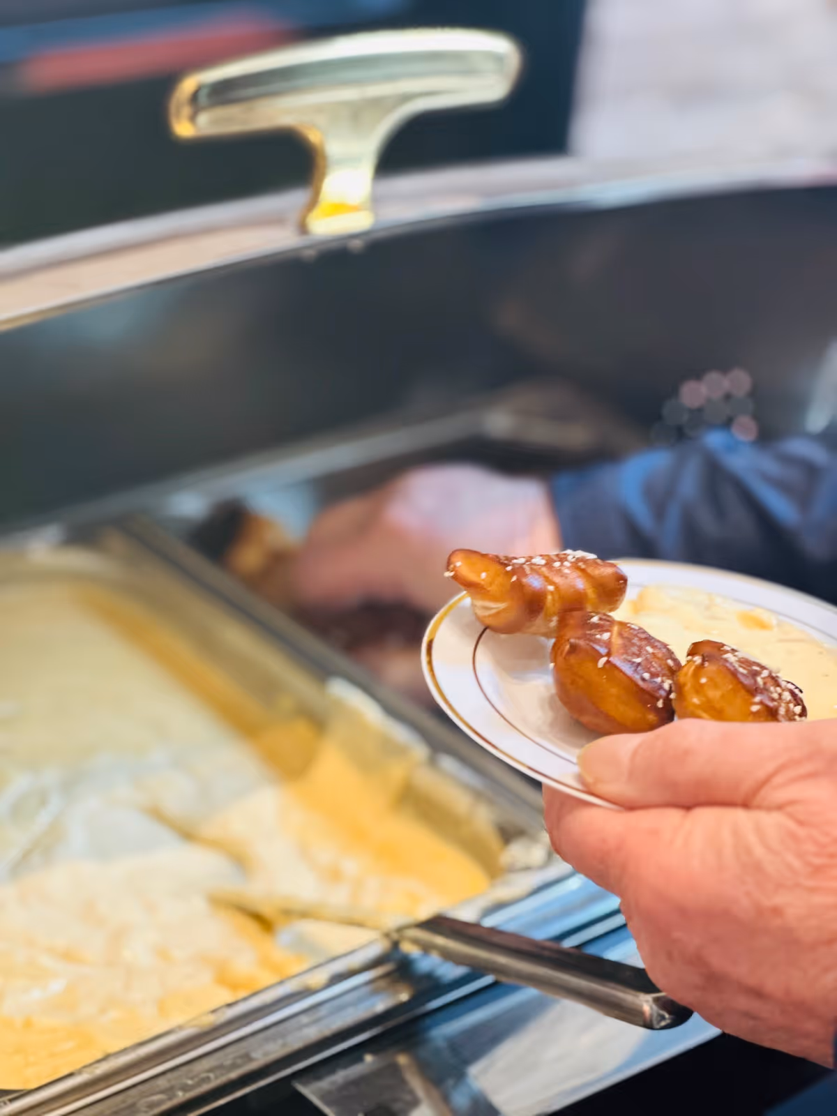 Close-up of a person holding a plate with soft pretzels and cheese sauce next to a buffet tray filled with melted cheese sauce.