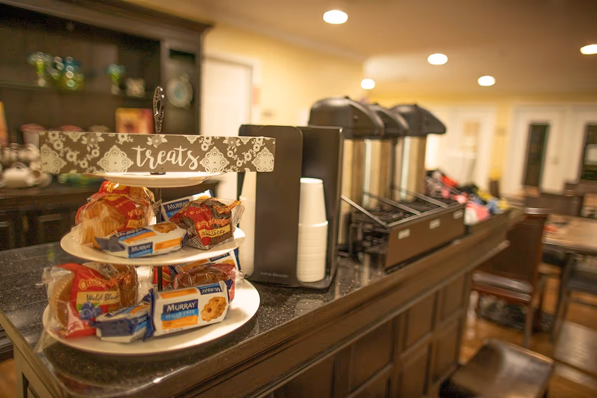A countertop with a three-tiered tray labeled 'treats' holding packaged snacks and muffins. Next to the tray are three large coffee dispensers with stacked disposable cups. The background shows a warm, softly lit interior with wooden furniture and shelves.