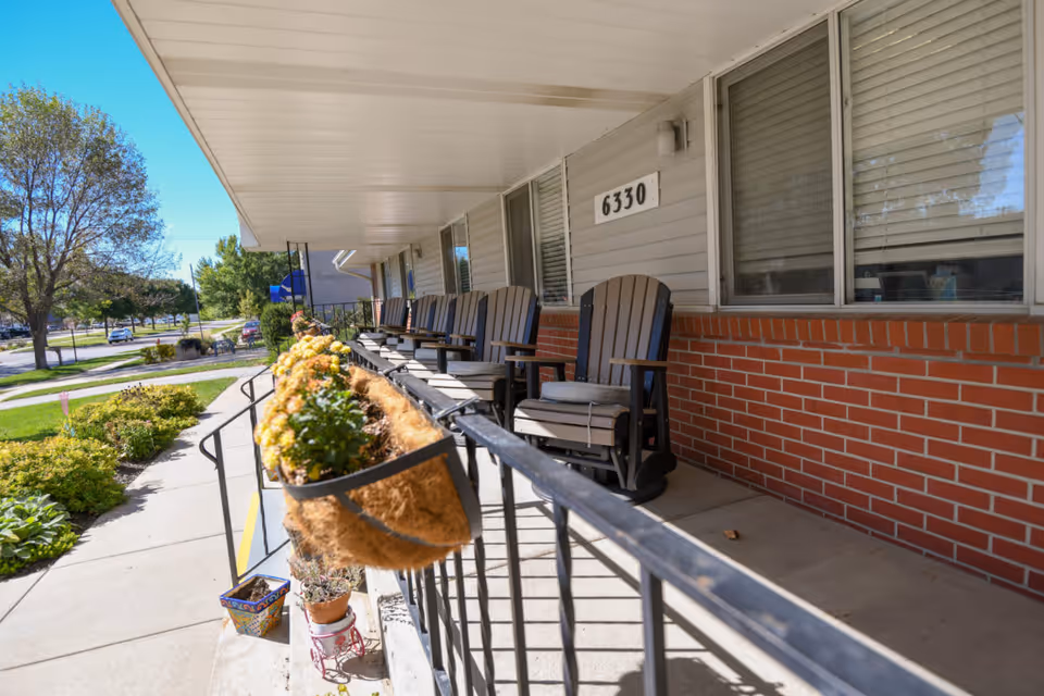Outdoor porch area of Havelock Manor featuring a row of wooden rocking chairs with cushions along a brick and siding exterior wall. There is a railing with flower planters attached, and a sidewalk with greenery and trees visible in the background under a clear blue sky.