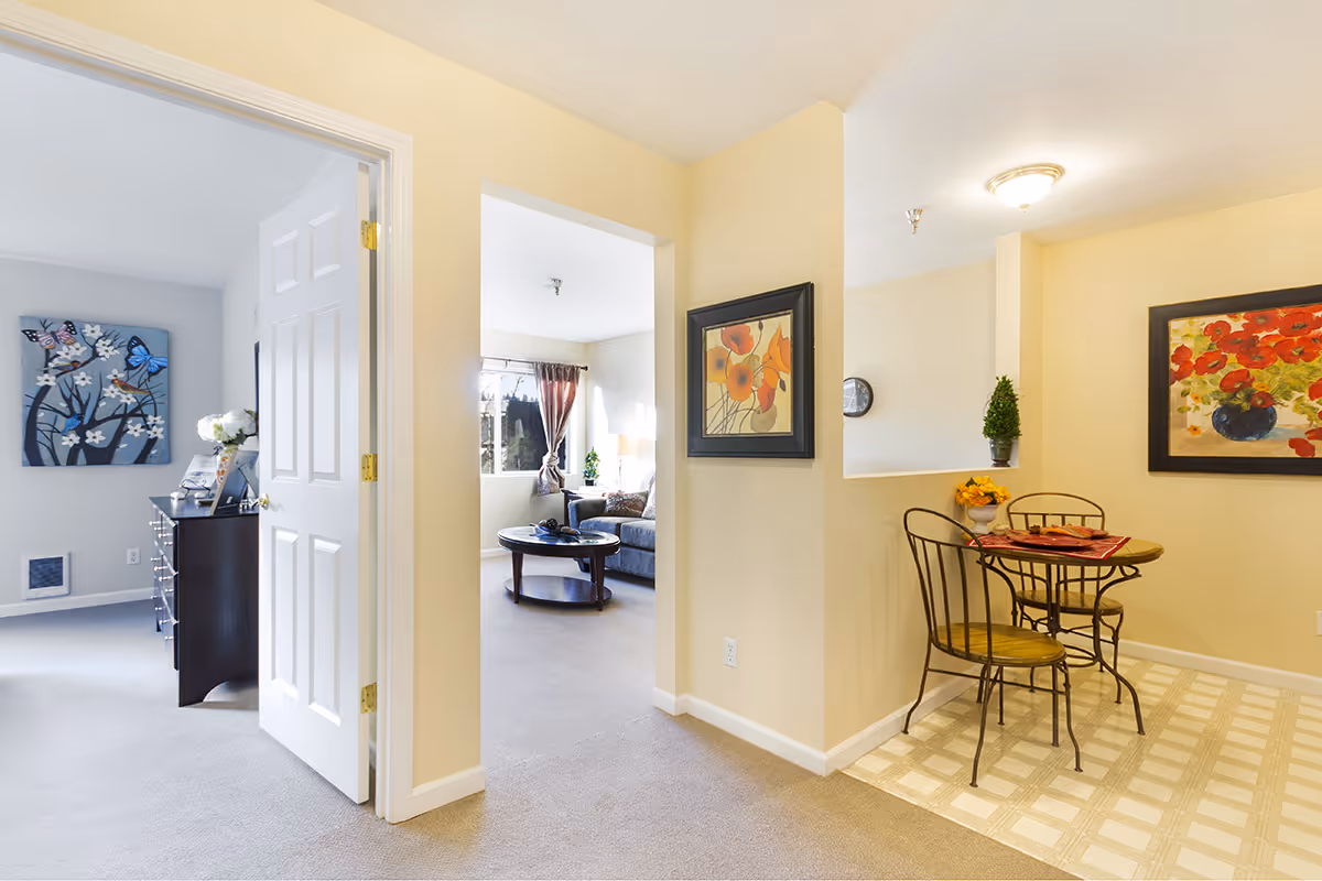 Interior view of a senior living facility showing a small dining area with a round table and two chairs on the right, decorated with a flower vase and wall art. To the left, there is an open doorway leading to a bedroom with a dresser and butterfly-themed artwork on the wall. In the center, an open passage leads to a living room with a coffee table, sofa, and window with curtains letting in natural light.