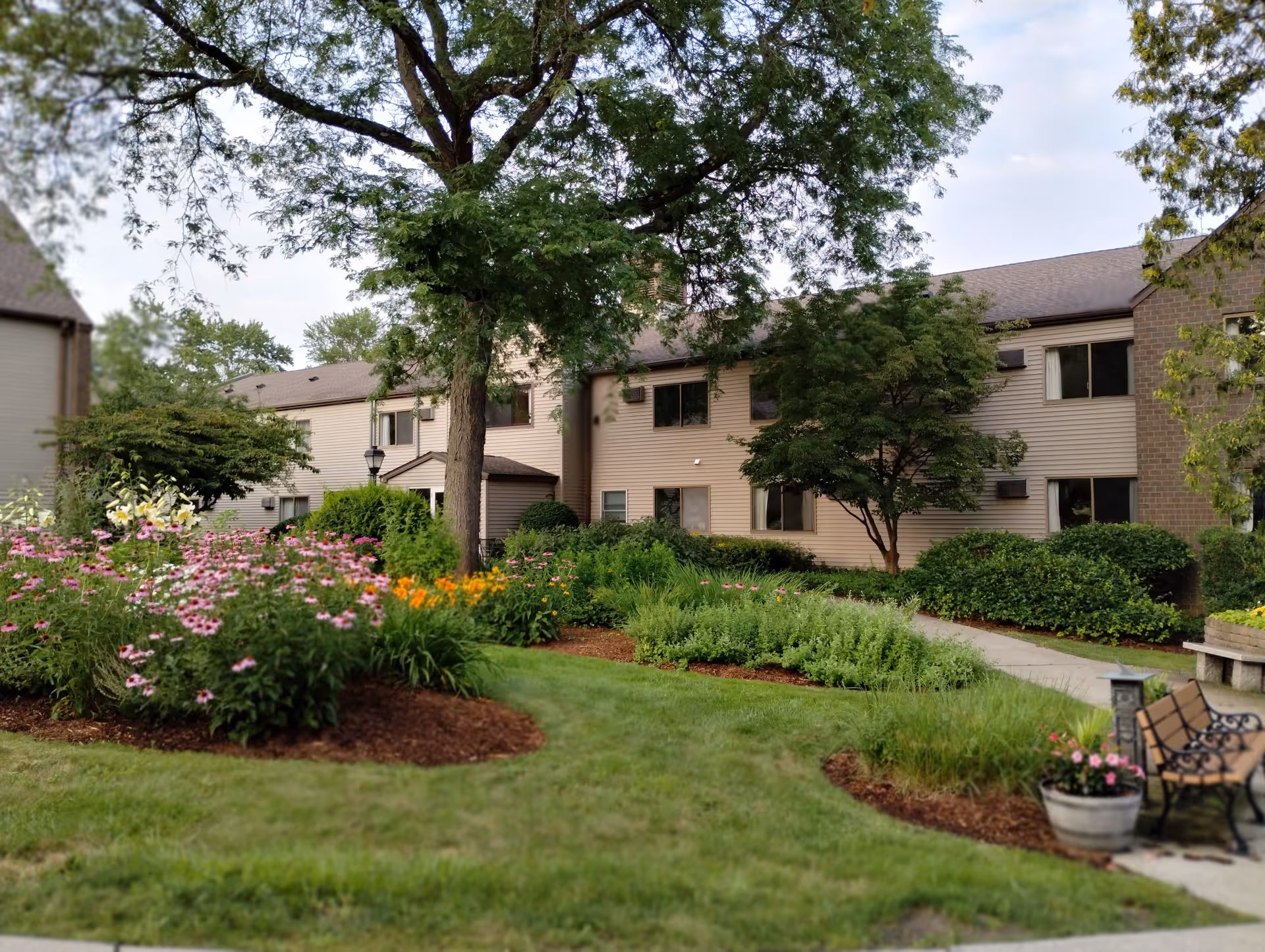 A landscaped garden area with colorful flowers, green shrubs, and trees in front of a two-story senior living facility building. There is a paved walkway, a bench, and a planter with flowers near the path.