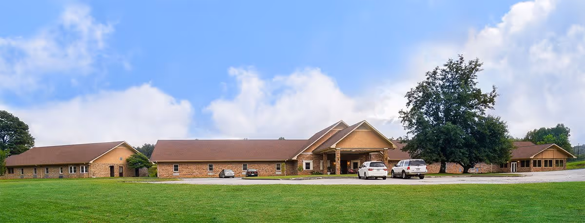 Wide exterior view of a single-story brick building with a brown roof, surrounded by green grass and a few trees under a partly cloudy sky. Several cars are parked near the entrance.